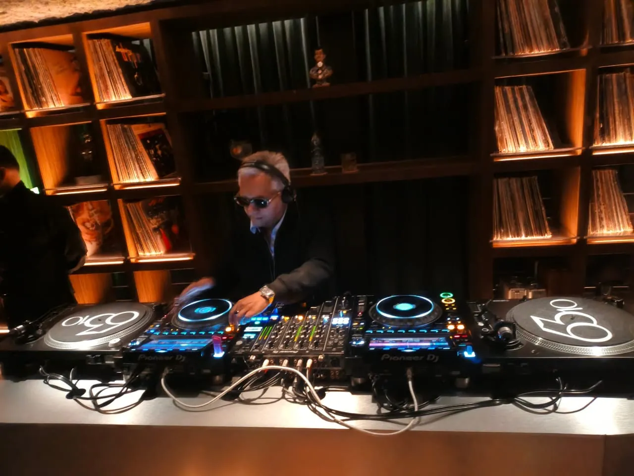 Charles Leiv mezclando en una cabina con dos tornamesas y dos reproductores Pioneer, frente a estantes de vinilos