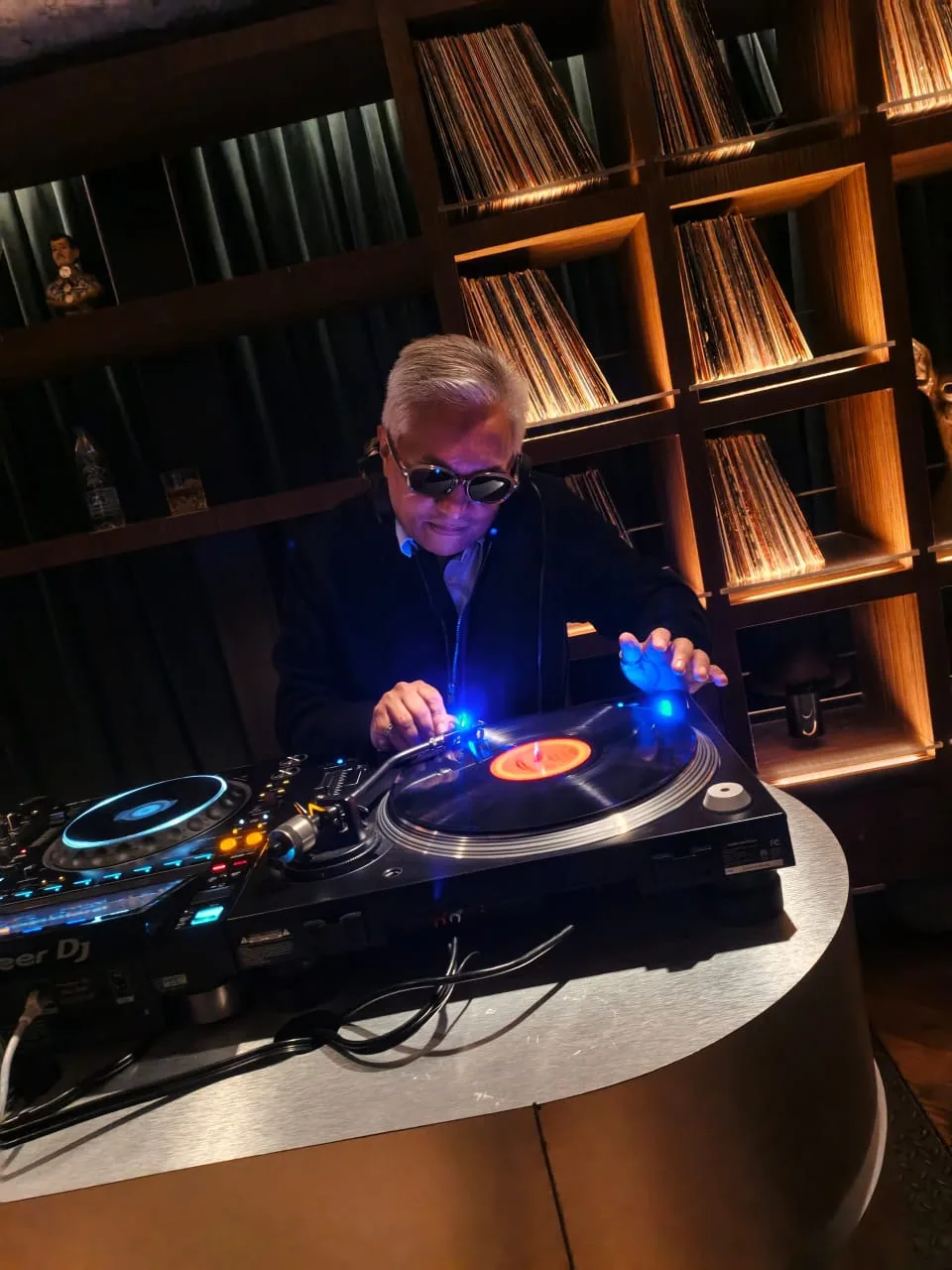 Charles Leiv con audífonos colocando un vinilo en el tornamesa, frente a una pared de discos iluminada